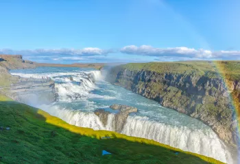 Wide shot of alpine vista, waterfall, rainbow, cliffs on either side. 