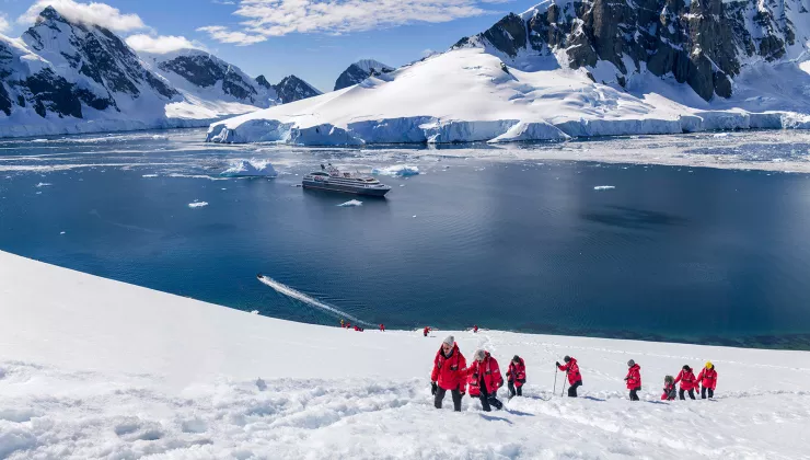 Hikers trekking up a snowy hill in Antarctica with a cruise ship anchored in the background
