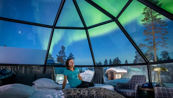 Woman sitting on a bed in a glass dome, with the Northern Lights in the sky