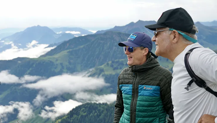 Two men wearing sunglasses on top of a mountain, looking towards a large, foggy valley