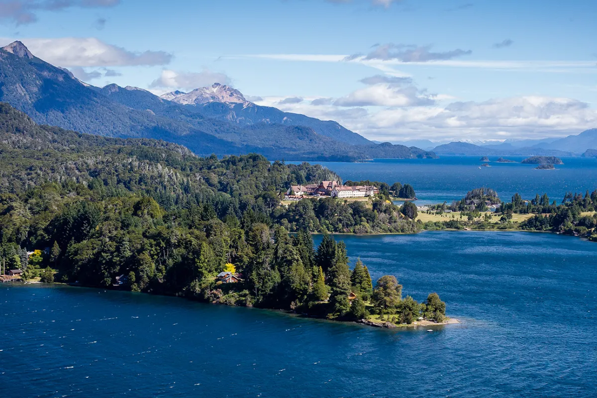Wide shot of inlet, white building, snowy peak, larger lake in distance.