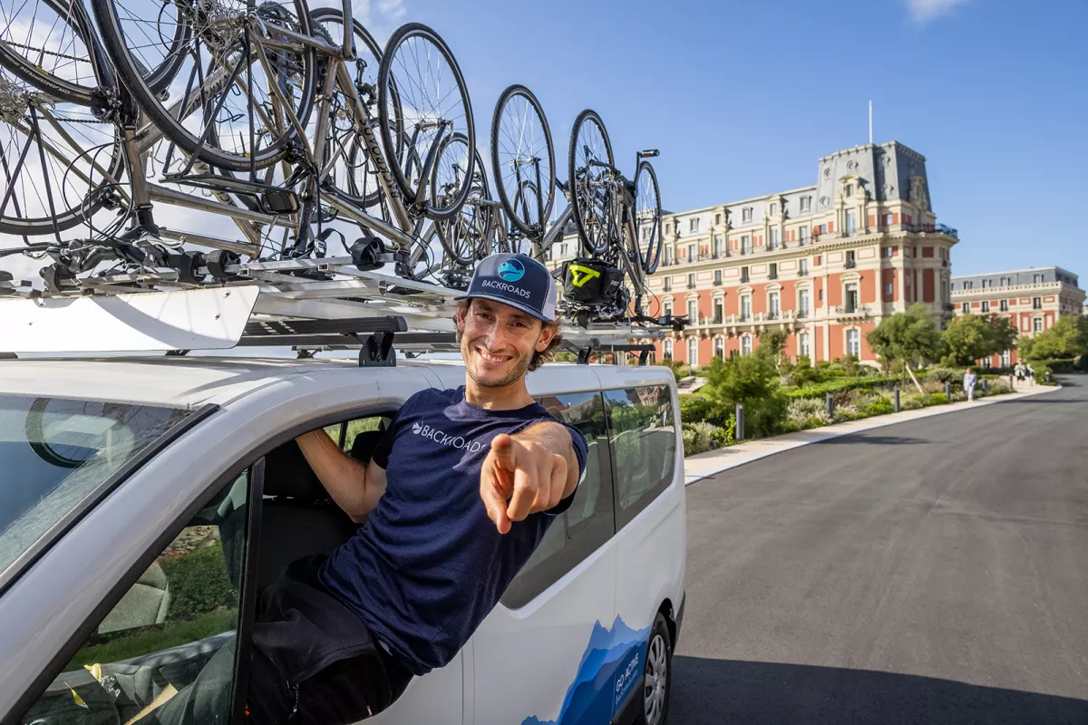 Leader sitting on van window, pointing to camera, Hôtel Du Palais Biarritz in background.