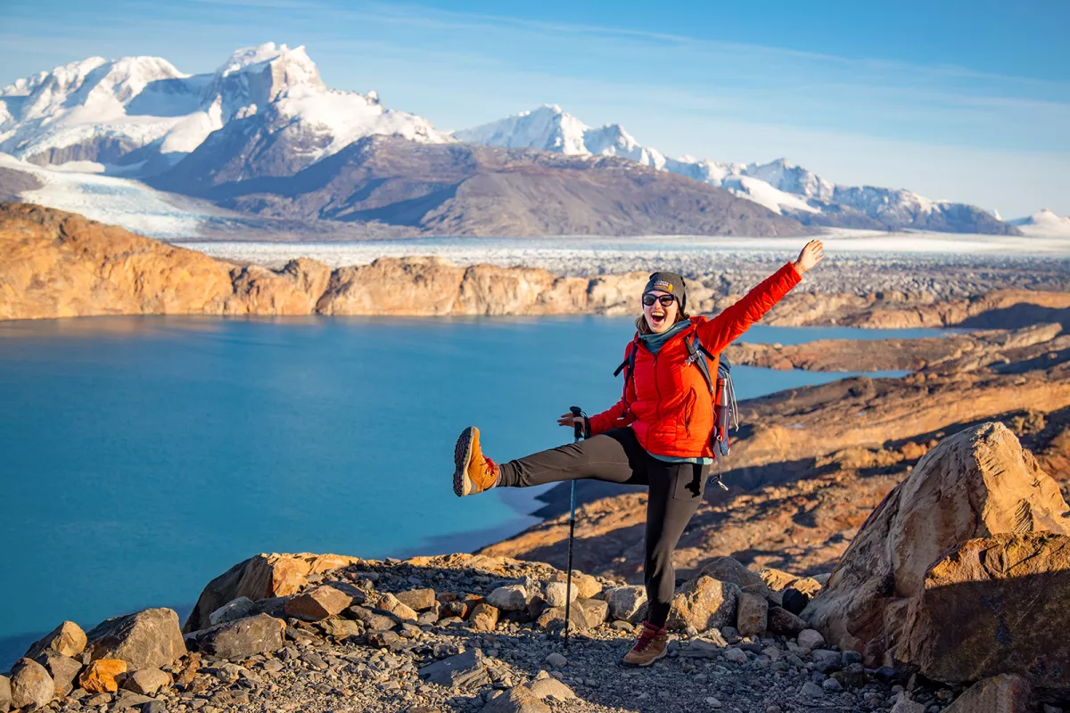 Woman in a red jacket with her arms wide open, walking on a rocky trail next to a lake