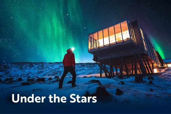 Dark figure admires the northern lights next to modern hotel building with text overlay "under the stars"