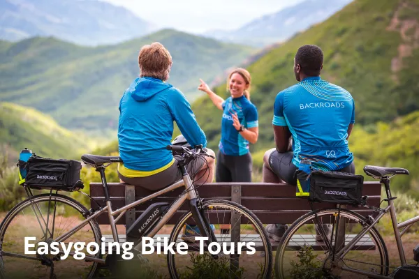 Three people in blue backroads jerseys sit while overlooking at a mountainous landscape. Easygoing Bike Tours is written in the bottom left corner.