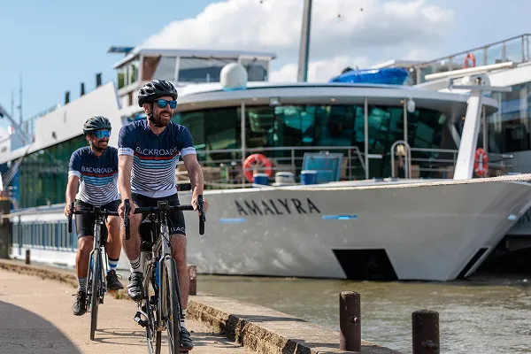 Two men biking alongside a river cruise ship in Francr