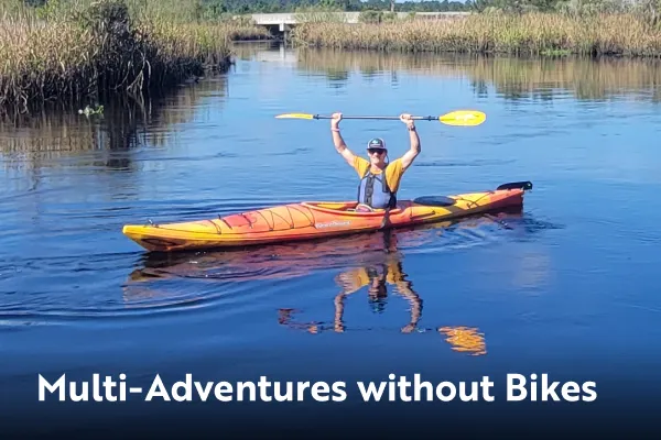 Man in a kayak raises paddle over his head. Multi-Adventures without bikes is written in the bottom left.