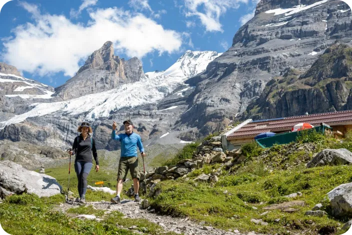 Two people hiking on a rocky and grassy trail, with large mountains in the distance