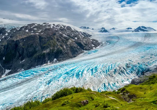 Shot of glacial vista, green cliff, blue ice, grey mountain.