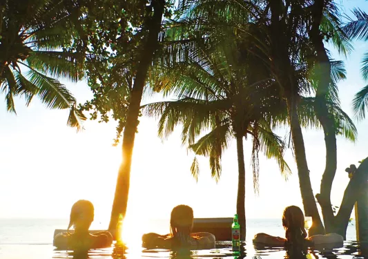 Three guests in pool, sun shining, palm trees above.