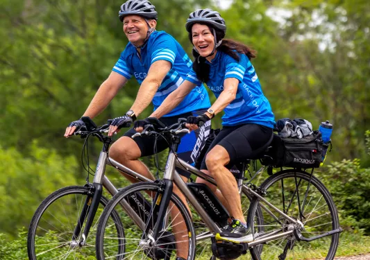 Two guests biking on forest road, smiling at camera.