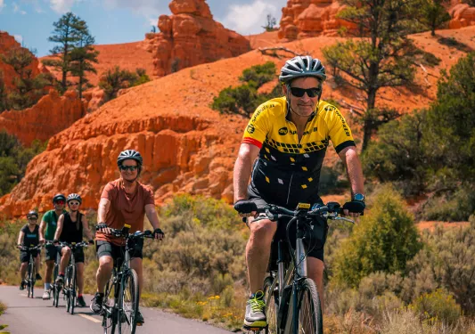 Five guests cycling towards camera, trees, vibrant red sand, hoodoos behind them.