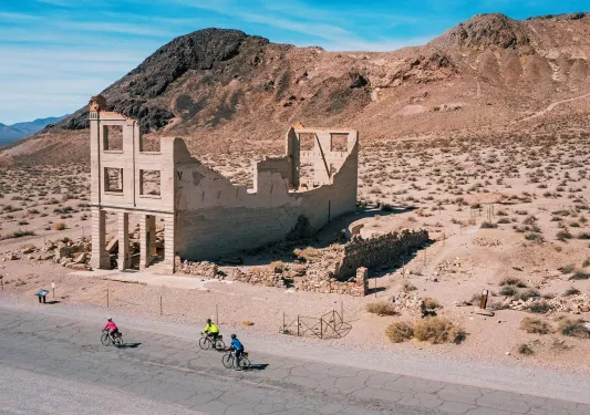 Guests biking through California desert with abandoned building in background.