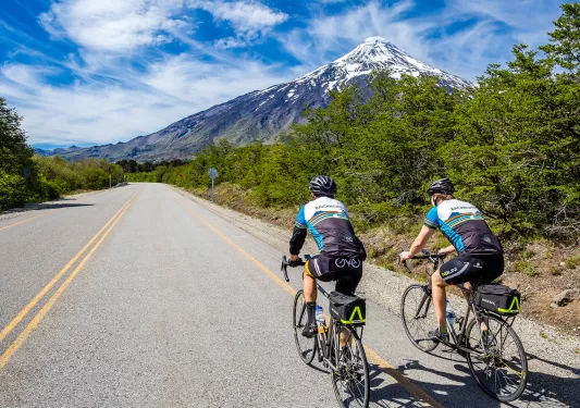 Two guests cycling down mountain road, snowy peak in background.