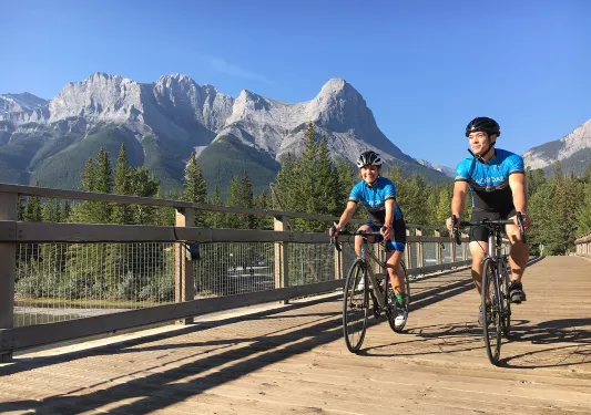 Two guests cycling on wooden path, Rockies in distance.