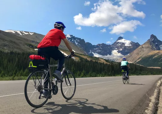 Two guests cycling down mountain road, snowy caps in background.