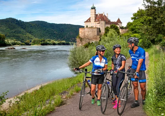 Three bikers walking their bikes on a path along the Danube River.