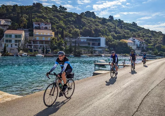 Four guests cycling down coastal road, blue ocean, hillside houses behind them.