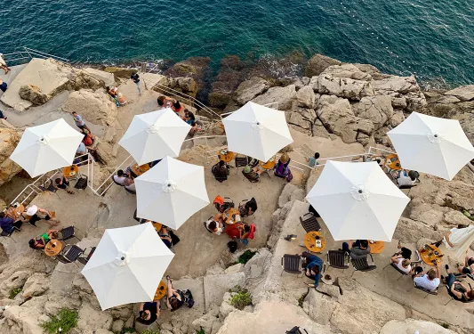 Overhead shot of people eating beneath white umbrellas, stone cliffs, ocean.