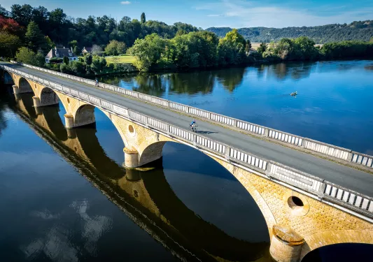 Two guests cycling on bridge, forest, meadow behind them.