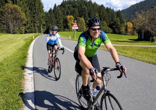 Two bikers riding down a hilly, tree lined street.