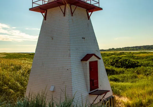 Small red and white lighthouse on a beach, sun peaking above.