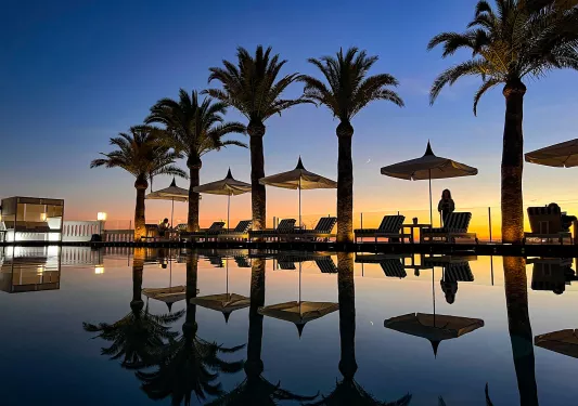Sunset shot of still resort pool, palms, recliners.