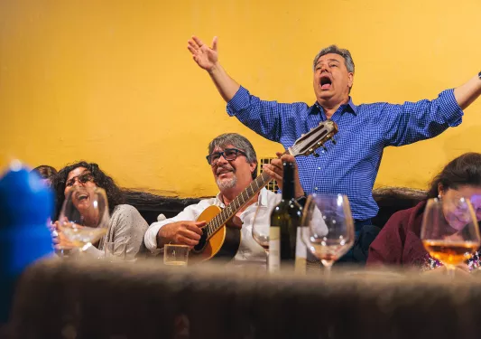 Guests at meal, one playing guitar, other singing behind them.