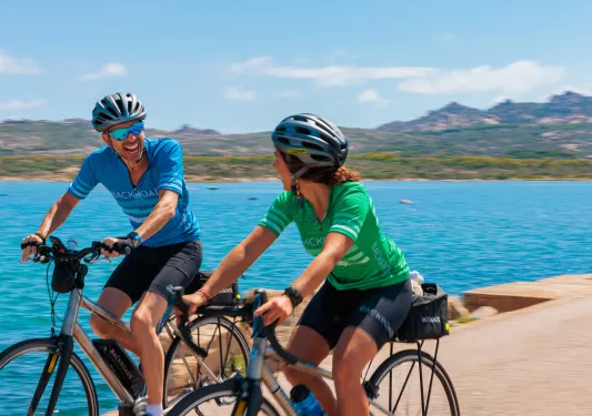 Two guests laughing and cycling, blue water, hills behind them.