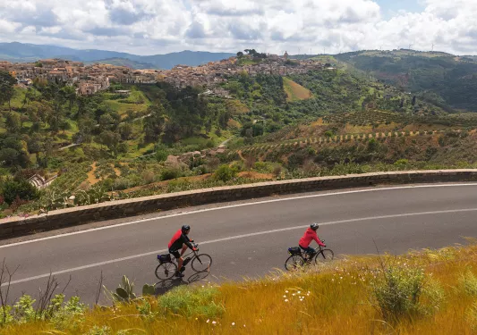 Two guests cycling down road, Medit. hilltop town in distance.