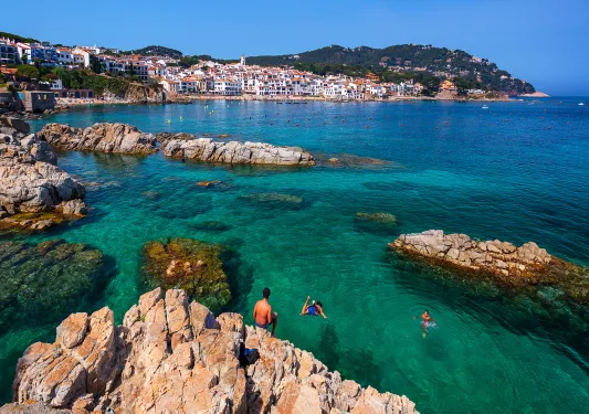 Guests jumping off craggy cliffs into blue water, Costa Brava in distance.