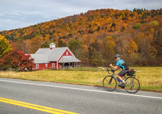 Guest cycling past red farmhouse. Fall trees behind.