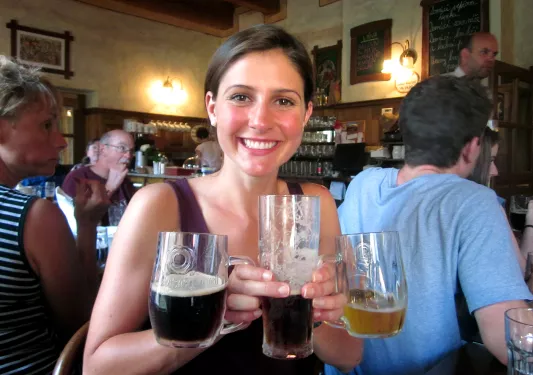 Woman posing in bar with three glasses of beer in varying colors.