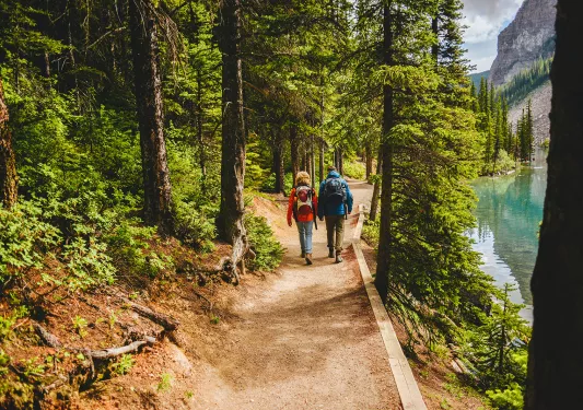 Two guests hiking down forest trail, river or lake to their right.