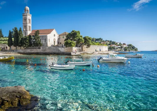 Wide shot of Hvar Island, white stone buildings, blue water, boats.