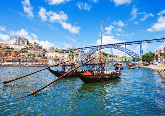 Two dark wood boats on Douro River, large metal bridge, colorful houses behind.