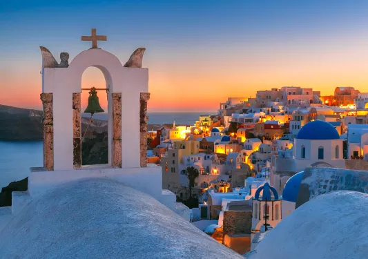 Sunset shot of Santorini, city lit up, church bell in foreground.