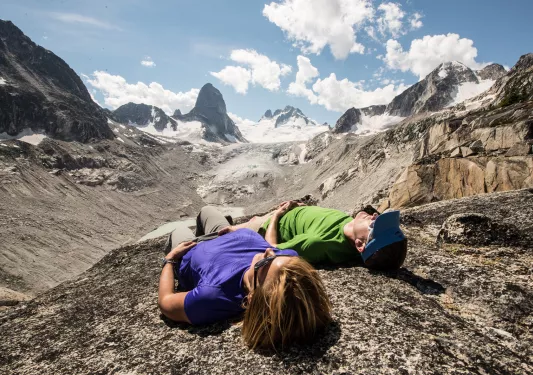 Two guests laying on rocky mountainside, arid cliffs and clouds in background.