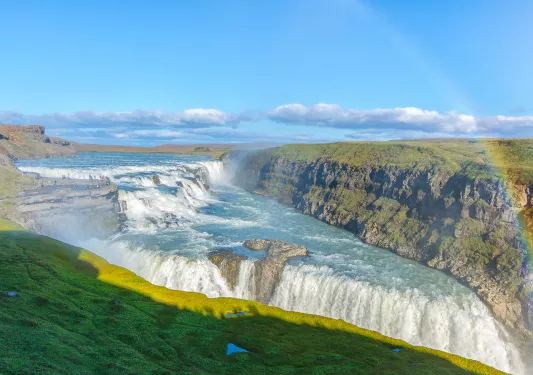 Wide shot of alpine vista, waterfall, rainbow, cliffs on either side. 