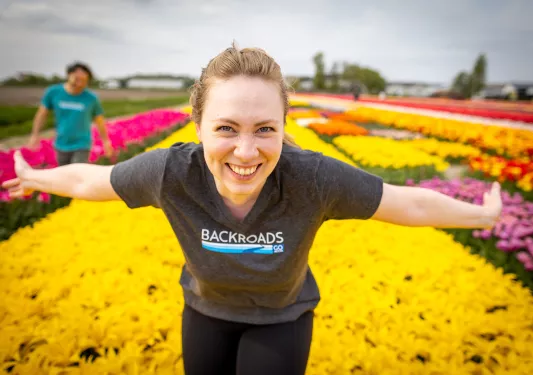 Guest among flower beds, gesturing to them, smiling into camera.