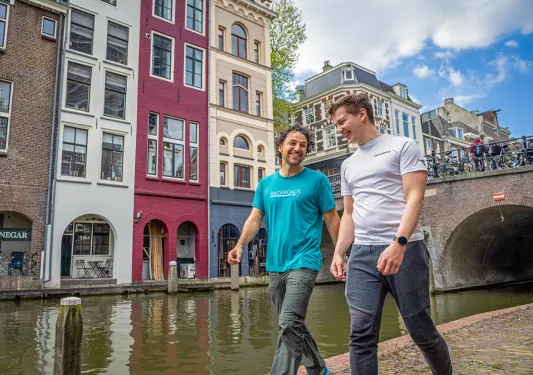 Two guests walking next to a river in town, colorful storefronts behind them.