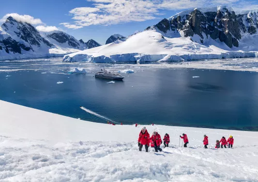 Hikers trekking up a snowy hill in Antarctica with a cruise ship anchored in the background