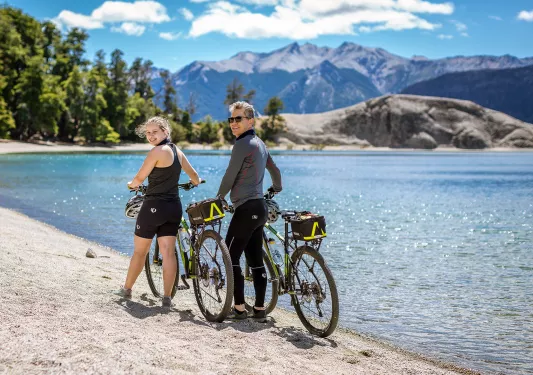 Two guests with bikes, walking along beach, blue water, forest behind them.