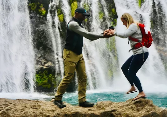 Two guests walking over rocks, waterfall behind them.