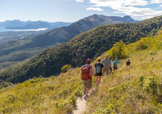 Group of guests walking down hillside, mountains, river in distance.