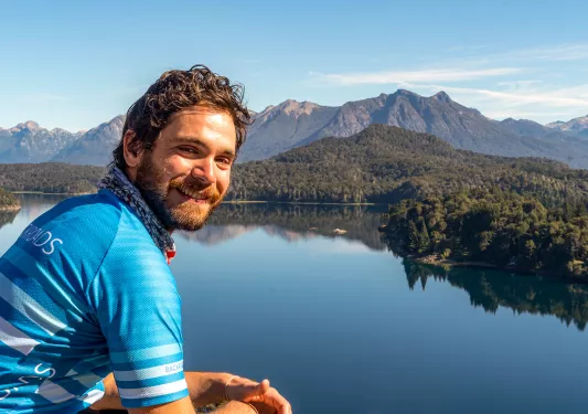 Guest posing next to still lake, forest, mountain behind them.