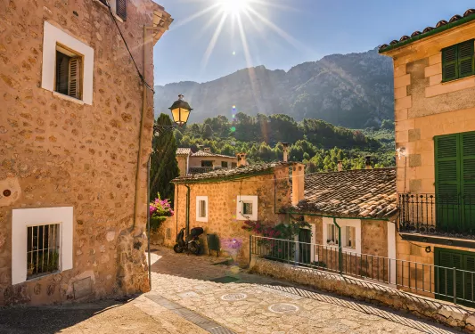POV shot in tan stone village, sun shining, forested mountains in distance.