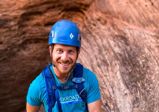 Guest in rock climbing gear on cliffside, smiling at camera.