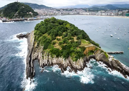 Bird's eye shot of craggy forested island, coast in distance.