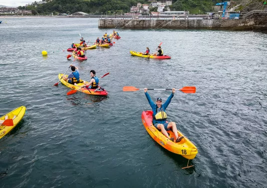 Group of guests in kayaks, one raising paddle over head, coast behind them.